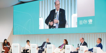 Six speakers on stage at WUF13. Second from the right is Habitat CEO Jonathan Reckford, who can also be seen on a large screen above the stage speaking into a microphone. 