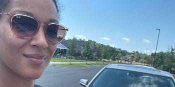 Up close of a woman in sunglasses and black shirt posting with her silver car.