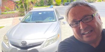 Man with gray hair, glasses and a dark blue shirt posing up close with his silver Toyota