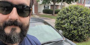 Close up selfie of a man in black sunglasses and a dark beard posing with his dark colored car. 