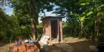 Photo of a small, brick structure, a latrine, near green trees.