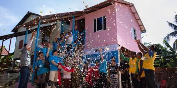 A group of volunteers celebrate in front of a newly built pink Habitat house by throwing confetti in the air.