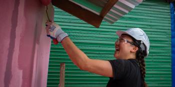 Volunteer in a hard hat paints a building pink with a paint roller.