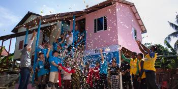 Habitat volunteers and community members celebrate a completed build in front of a pink house as they throw colorful confetti in the air.