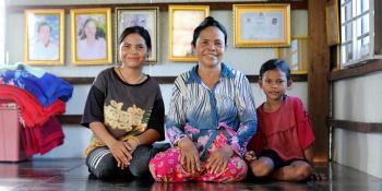 Rem is flanked by two of her granddaughters, seated on the floor in their home in Battambang, Cambodia. Behind them a wall of gold frames displays family photos and awards.