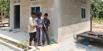 A man and boy on either side of an elderly woman, helping her walk down a ramp walkway out of her house.