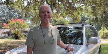 Bald man in a gray t-shirt with glasses hanging around his neck posing in front of his silver car.