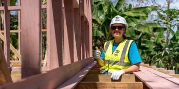 A woman in a bright blue T-shirt, hardhat and yellow vest gives a thumbs-up while standing between wooden slats of a Global Village house build. Palm trees are in the background.