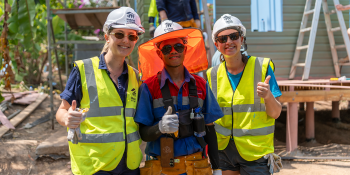 Three volunteers wearing hardhats, sunglasses and safety equipment pose together in front of a house under construction and palm trees.