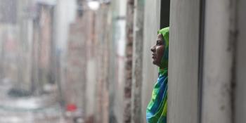 Bangladeshi woman in green looks out the doorway of her concrete home during heavy rain