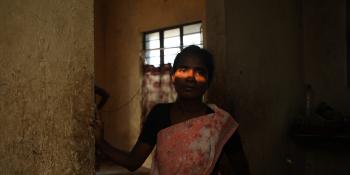 A woman in a sari stands in a doorway. Her hand is on the wall and a swath of light from the window illuminates her eyes.