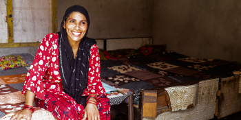 A woman sits in her home wearing a black head scarf and red shirt and pants, smiling from the camera.