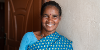 A woman stands smiling and wearing a blue sari with polka dots in front of a white wall and brown door.