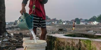 A person in a red shirt draws water from a community well using a broken blue bucket.