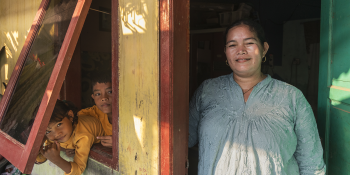 A mother stands in an open front door while her children lean out of a window, all staring at the camera.