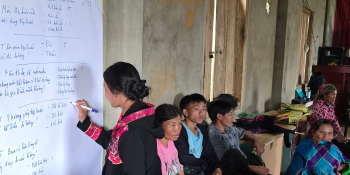 Woman writing on a large piece of paper on a wall. Several other people are seated behind her. 