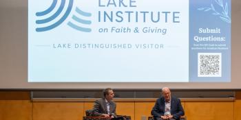Jonathan Reckford and another man sitting onstage in chairs in front of an audience. In the background, a projector screen displays "Lake Institute on Faith & Giving" "Lake Distinguished Visitor" and a QR code. 