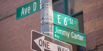 Street sign pole. Green street sign at top left reads "Ave D." On the right, green street signs read "E 6 St" and "Jimmy Carter Way." Below the other signs, a black and white arrow sign reads "ONE WAY."."