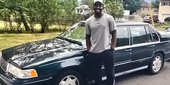 Man in gray shirt, black pants, tennis shoes and a white hat posing in front of a dark colored car.