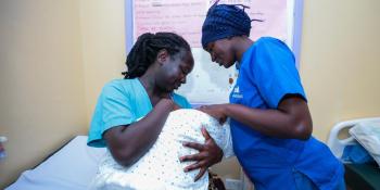 Kenyan nurse dressed in blue scrubs checking on a Kenyan mother and her new baby in a clinic.