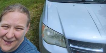 Woman with blond hair in a blue t-shirt posing with a silver car.