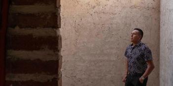 A man wearing a gray shirt and black jeans stands in profile staring up at a wall.