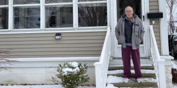 An older man in a jacket stands on the porch of his white house, surrounded by snow.