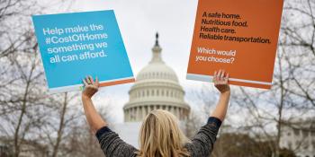 Una mujer rubia, de espaldas a la cámara, mira hacia el Capitolio en Washington D.C. y sostiene un cartel azul y otro naranja en defensa de la vivienda.