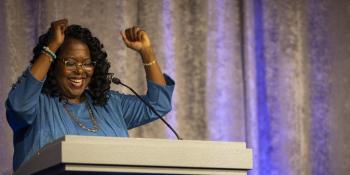 Black woman in a blue blouse celebrating with her hands in the air at a podium with HFHI's logo. 