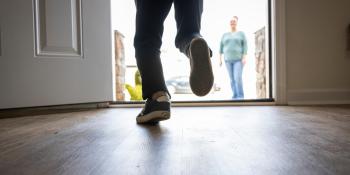 A child's legs run out past an open white front door toward his mother who is standing and smiling in the front yard.