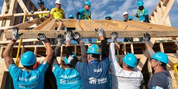 A group of volunteers work together to lift up the frame of a house while more volunteers stand on top of the roof pulling the frame up.