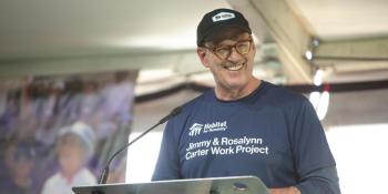 Jonathan Reckford stands at a podium smiling and wearing a Habitat shirt and black baseball hat.  