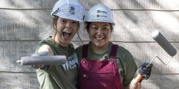 Two college volunteers in hard hats smile while holding paint rollers on build site.