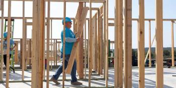 A volunteer in a blue hard hat and Habitat shirt walks through the wooden frame of a house build holding an armful of lumber.