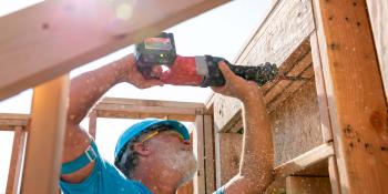 A volunteer stands with his arms above his head holding a drill that he is pushing into the frame of a house.