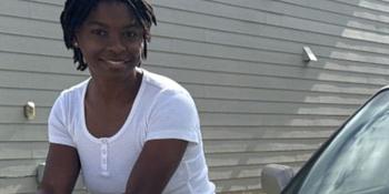Woman in white t-shirt and tan shorts sitting on the hood of a tan car.