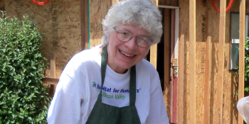 Ellen Hill smiles at the camera with house framing on a Habitat build site in the background. She has short white curly hair, glasses, and is wearing a Habitat shirt and green apron.
