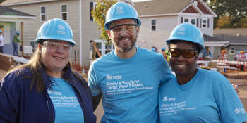 Two female volunteers and one male volunteer pose together smiling with a house construction site in the background. They are wearing light blue hardhats and matching blue T-shirts branded with the Habitat logo and "Jimmy & Rosalynn Carter Work Project"