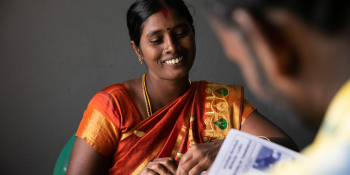Indian woman in bright orange clothing sits across a table from a man looking at paperwork.