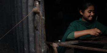 Young girl in green clothing smiling as she weaves on a loom inside her home