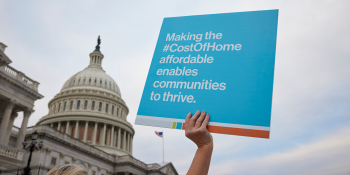 Hand holding sign that says "Making the Cost of Home affordable enables communities to thrive" with U.S. Capitol building in background
