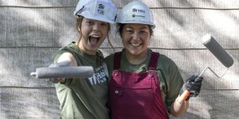 Two young women volunteers with white hard hats and paint rollers smile while posing together against the side of a house.
