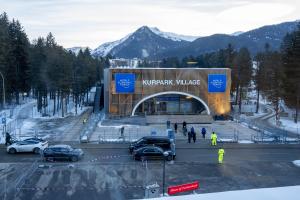 High angle photo showing the outside of the World Economic Forum venue surrounded by trees, with the Swiss Alps mountains visible in the background. The building sign says "Kurpark Village" in white text, and a blue banner is hung on either side with the World Economic Forum logo.