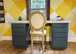 Blue desk with light wood top in front of a yellow wall with a chair in front. 