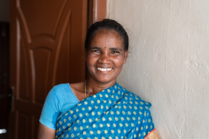 A woman stands smiling and wearing a blue sari with polka dots in front of a white wall and brown door.