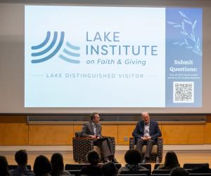 Jonathan Reckford and another man sitting onstage in chairs in front of an audience. In the background, a projector screen displays "Lake Institute on Faith & Giving" "Lake Distinguished Visitor" and a QR code. 