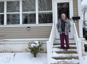 An older man in a jacket stands on the porch of his white house, surrounded by snow.