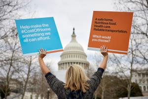 Una mujer rubia, de espaldas a la cámara, mira hacia el Capitolio en Washington D.C. y sostiene un cartel azul y otro naranja en defensa de la vivienda.