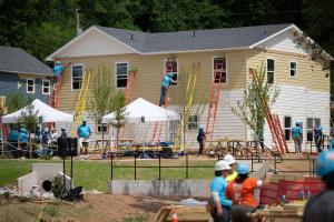 Volunteers working on the site of the 2026 Carter Work Project in the midground. In the background, a multi-family home is being painted by volunteers on ladders, as other volunteers work below. 