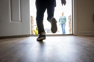 A child's legs run out past an open white front door toward his mother who is standing and smiling in the front yard.